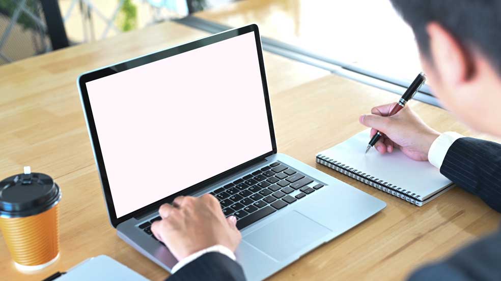 Personne prenant des notes sur un carnet tout en regardant son ordinateur allumé